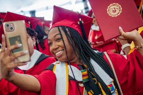 Woman is taking a selfie of herself in a red cap and gown at commencement.