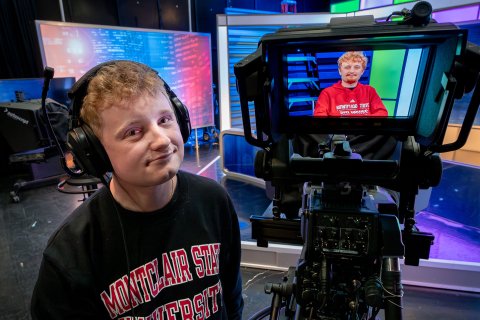 Brian Scott operates a professional television camera in a studio, wearing headphones and a Montclair State University sweatshirt. The camera’s monitor displays his brother, Craig Scott, who is seated on set and visible on the viewfinder, also wearing Montclair apparel.