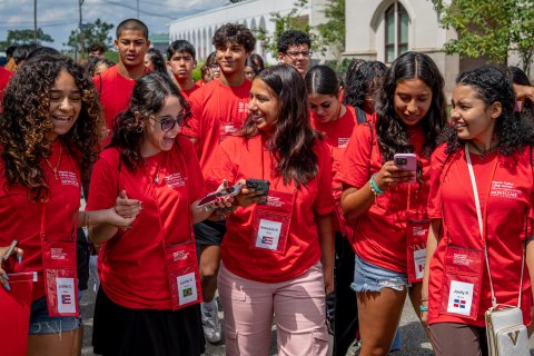 A group of students in red t-shirts and lanyards walk together in a large group on college campus.