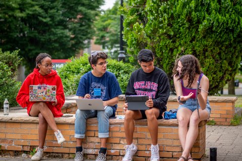Four students with laptops talk while sitting on a a low wall.