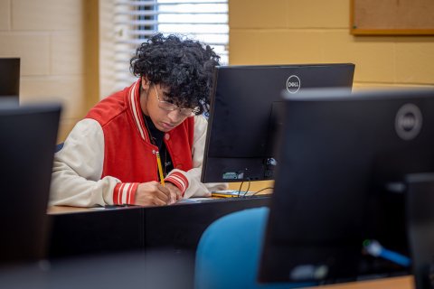 Man in a red letter jacket in front of a computer writing down information.