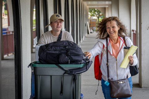 Alt Text: A parent and student push a cart loaded with college essentials into a dorm room.