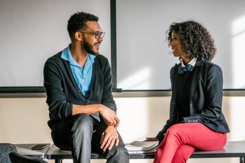Two students talk to each other while seated on a table.