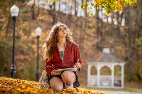 Student holds a pencil and pad while sitting on Montclair's campus. The fall foliage is orange and. yellow.