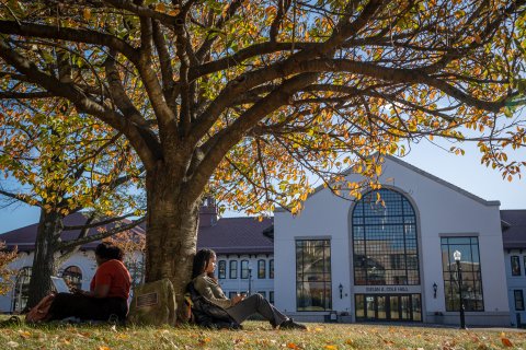 Two students sit under a tree with Cole Hall in the background.