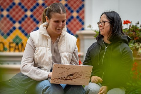 Two students are sitting in an outdoor courtyard, smiling and working on a laptop.