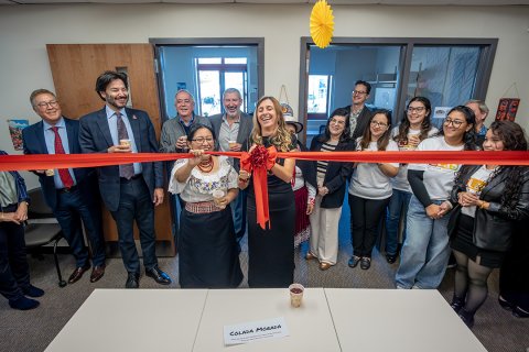 A smiling woman in a black dress cuts a ceremonial red ribbon in a classroom as spectators look on.