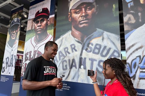A young reporter interviews a New Jersey Jackals baseball player in front of a display featuring historical baseball players at the Hinchliffe Stadium exhibit.