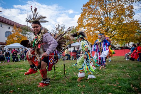 Native American men and women in traditional costumes dance outdoors while spectators watch.