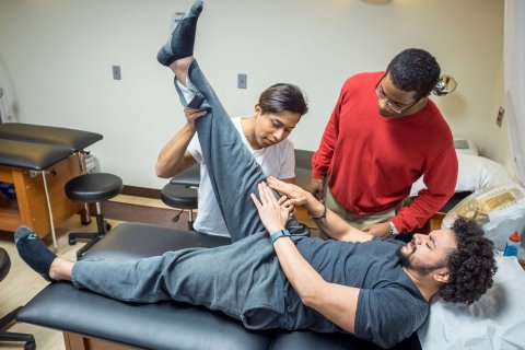 One student laying on a table describes how he's feeling to a trainer as the trainer stretches the student's hamstring. Another student looks on, observing the trainer.