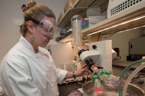 Student in chef's jacket wearing safety goggles working in the food lab.