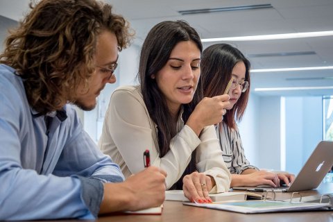 Two students in conversation at a table while a third student works on a laptop.