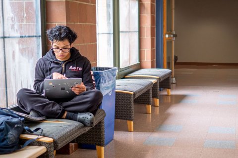 Student is sitting on a bench inside a building using his laptop.