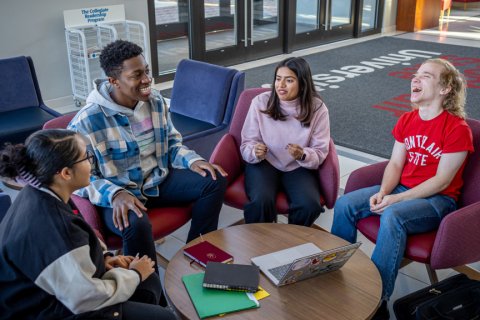 Group of students are sat in front of a round table conversating and laughing with one another.