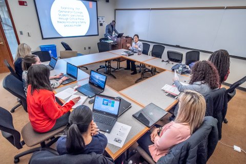 Student in a square shape desk wise at their laptops listening to their professor give a lecture.