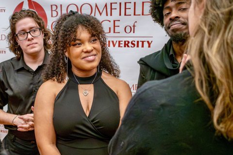 Ciara Donaldson, a 2024 Bloomfield College of Montclair State University graduate, stands at the center of the photo wearing a sleeveless black dress and a heart-shaped necklace, engaged with people gathered around her. Other individuals stand nearby, and a Bloomfield College of Montclair State University backdrop fills the background.