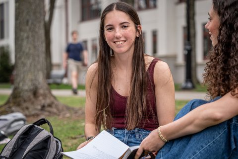 Dana Brzezinski sits on the grass at Montclair State University, holding an open notebook and talking with a friend, with campus buildings and a tree visible in the background.