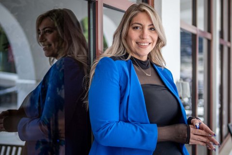 Ashley Crawford, in a black dress with bright blue blazer, stands in front of a building’s windows.