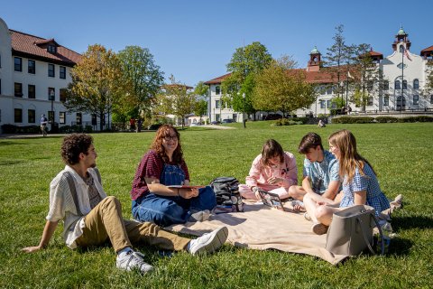 Students sit together on a blanket on a large grassy lawn at Montclair State University, talking and studying with notebooks and a laptop open, while academic buildings with red-tiled roofs and a clear blue sky frame the background.