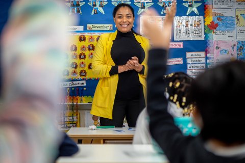 Teacher in yellow blazer at front of classroom facing young students