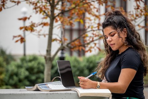 Female student at a table writing down notes in front of a laptop.