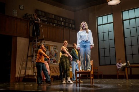 Four students on stage during a production. One student is standing on a chair in a spotlight.