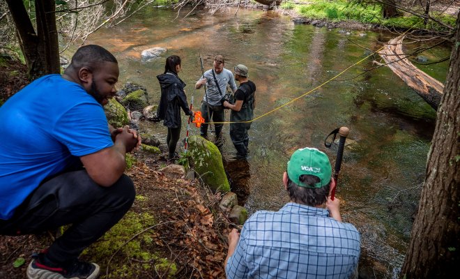 Two students perched on a river bank converse while watching three fellow students taking measurements in the water.