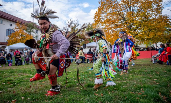 A group of Indigenous dancers dressed in traditional regalia adorned with feathers, beads and intricate patterns, perform outdoors on a grassy area during a cultural event. 