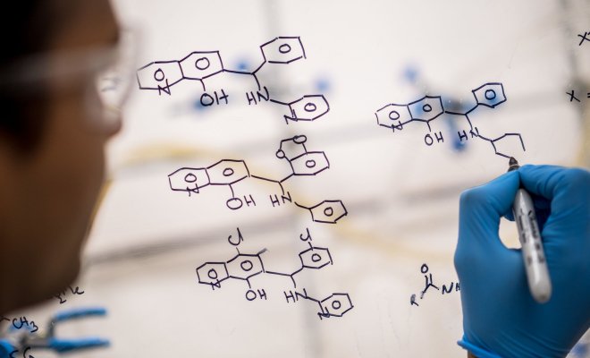 Photo over the shoulder of a chemist outlining a chemical structure with marker on a pane of glass