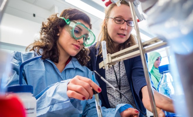 Professor looks over student's shoulder as she handles chemicals at a workbench using a pipette