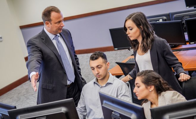 Three students and a professor all in businesswear collaborate on a project unseen behind a multiple-monitor workstation