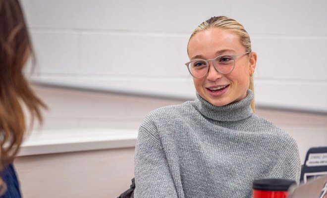 student in grey sweater and glasses speaking with someone in the foreground wearing a blue dress.