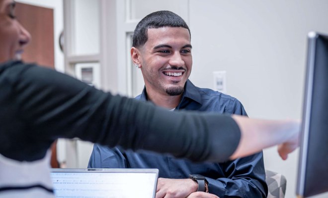 Two students smile as they look at a computer monitor.