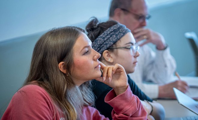 Three students sit in a classroom; two are listening and one is taking notes.