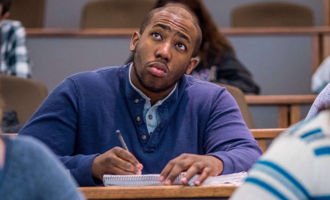 A student takes notes in a lecture hall with other students in the foreground and background.