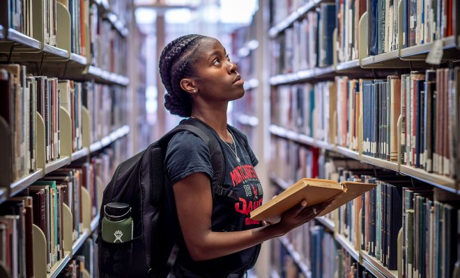 Person standing in library aisle holding an open book.