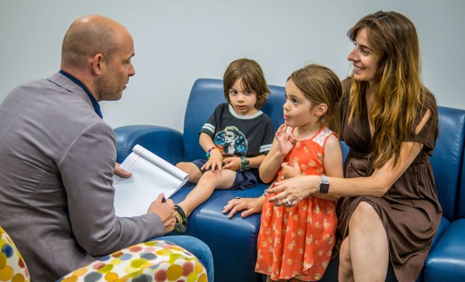 A seated man with a notepad speaks to a seated, smiling woman and a young girl as a young boy looks on.