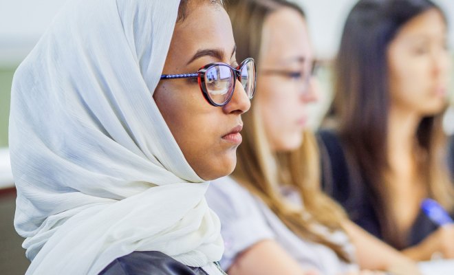 A woman wearing a white hijab sits, listening, in a classroom while two other women sit, listening, in the background.