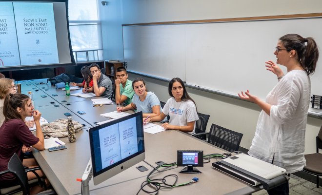 professor speaking to students gathered around a table with projection screen in the background.