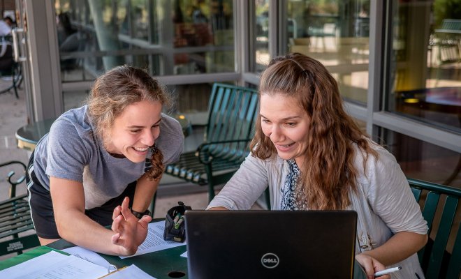 Two students talking over a laptop, notebook in the foreground with French translations.