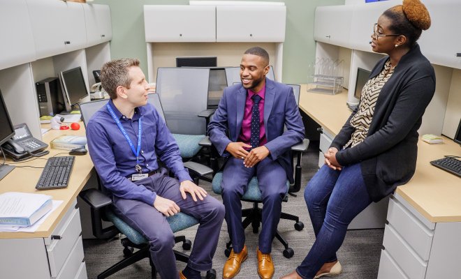 Two graduate students and one professor in professional attire have a discussion in an office space.