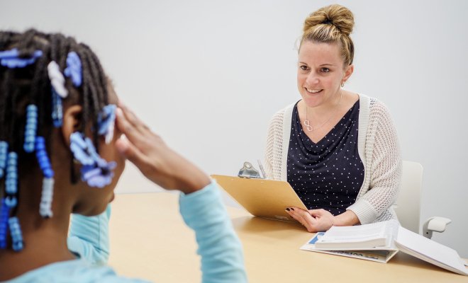 A young girl with braided hair and decorative beads sits at a table, facing an adult woman who is holding a clipboard and some papers.