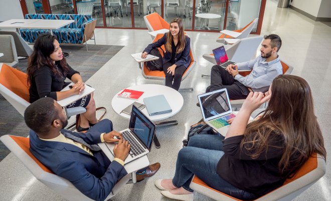 Five people are seated in a modern lounge area, around a small white table. Each person sits in an orange chair and is engaged with laptops, notebooks, or paper folders.
