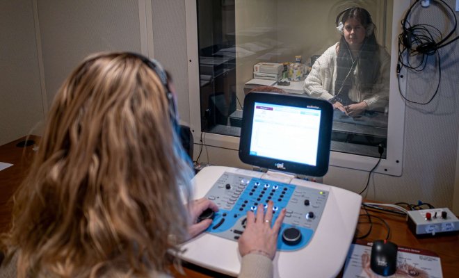 Student at a control panel observing a person wearing headphones through window.