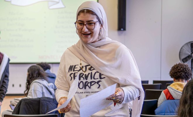 Student wearing hijab and a t-shirt that reads "next gen service corps" holding papers and speaking to another person.