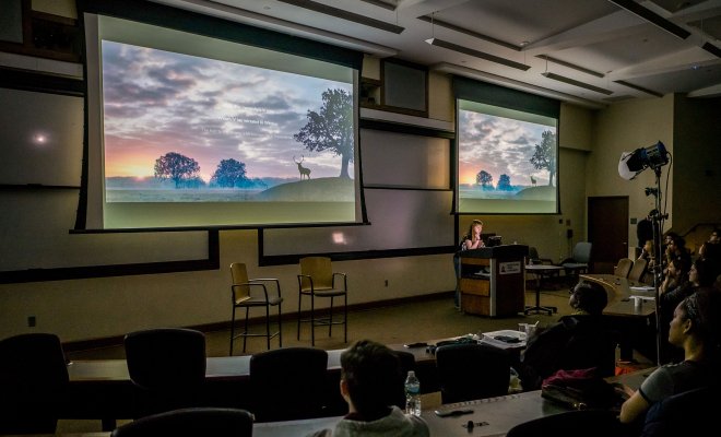 Students watch a film on projector screens in a darkened lecture hall.