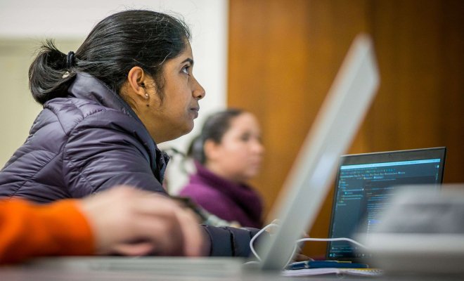 Profile of a woman looking up at the professor's screen as her laptop sits to the side with a screen full of computer code.