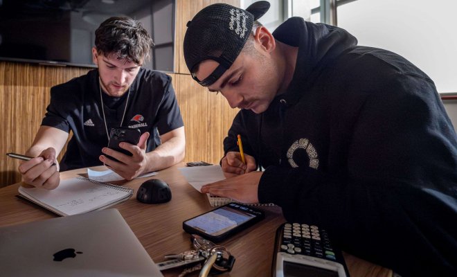 Two male students in lounge use graphing calculators while poring over notes.