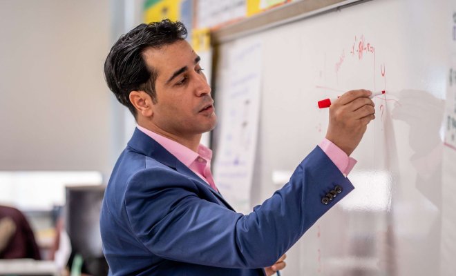 A teacher in a classroom stands before a whiteboard and writes out mathematics notes.