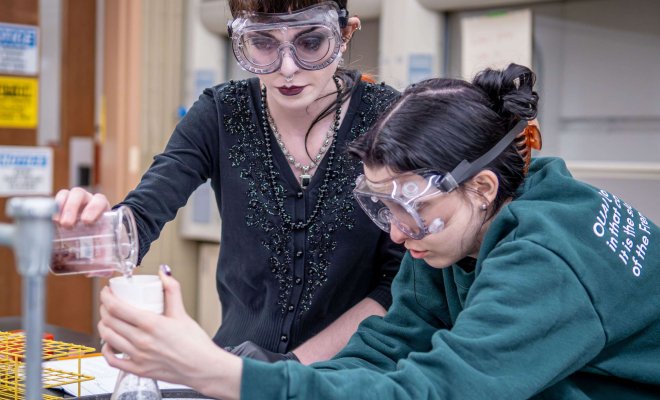 One student pours a liquid solution from a beaker while another student holds a funnel atop an Erlenmeyer flask.